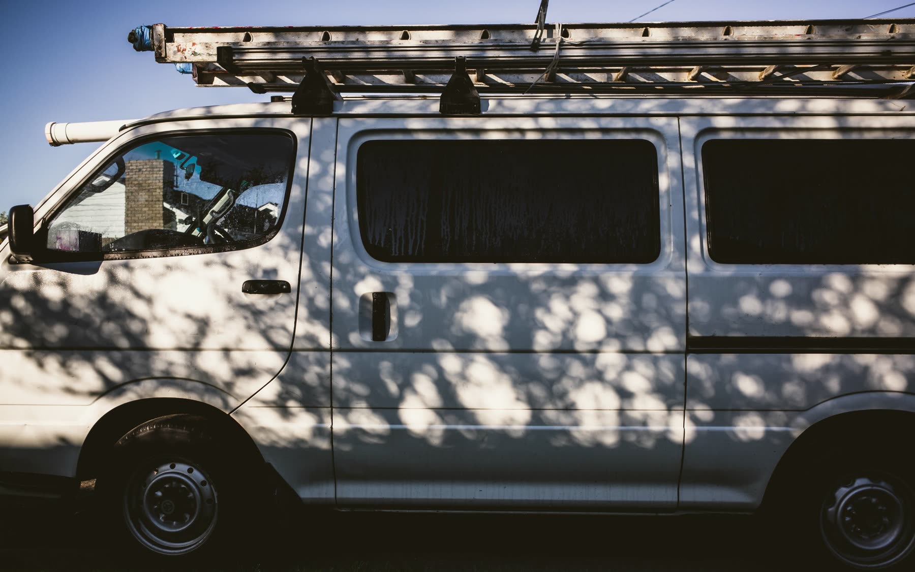 A white service van with a ladder on its roof rack, parked in dappled shade